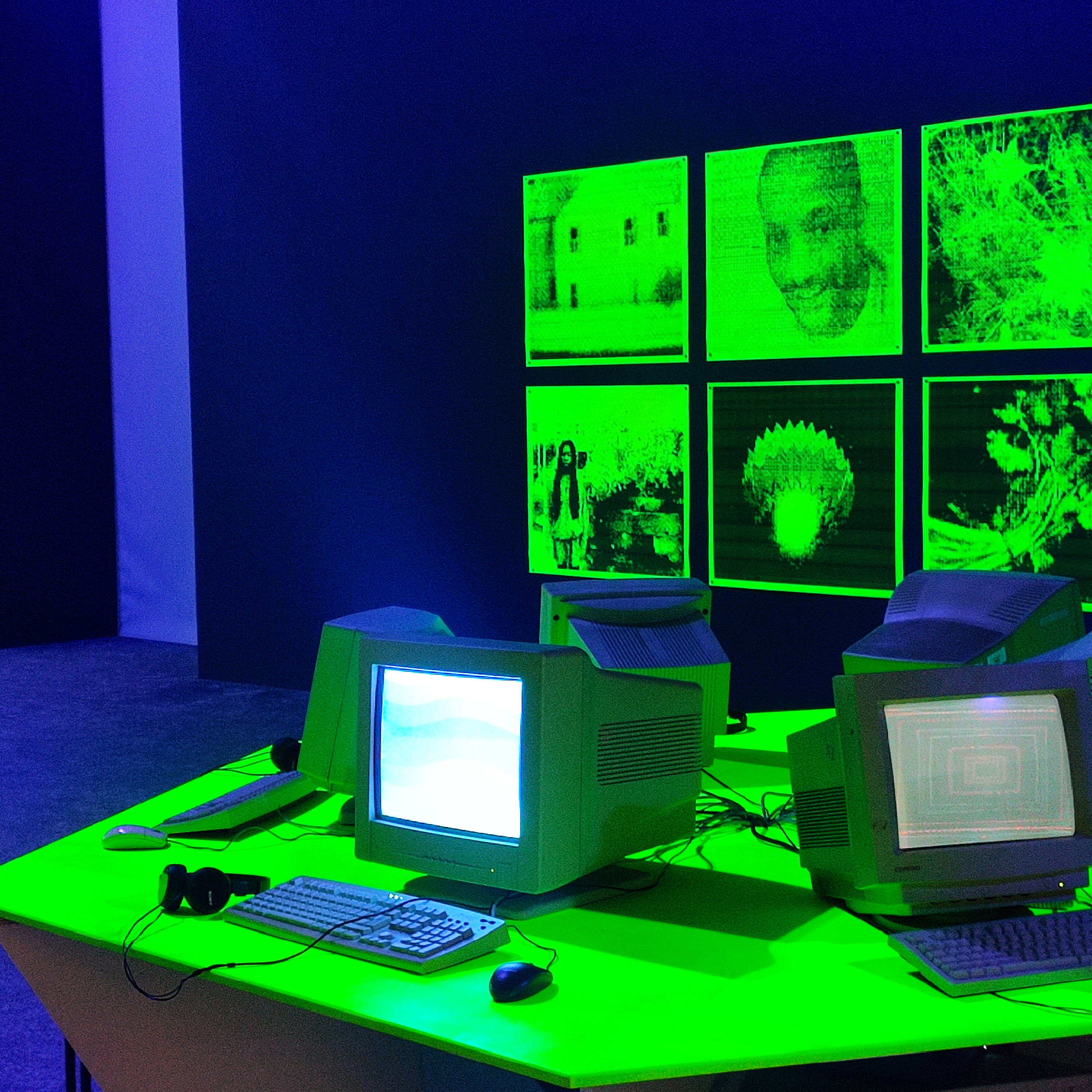 Part of the exhibition room with a dark backdrop, a neon green round table of retro computers with keyboards and headsets and in the background, there's a wall with neon green artwork.