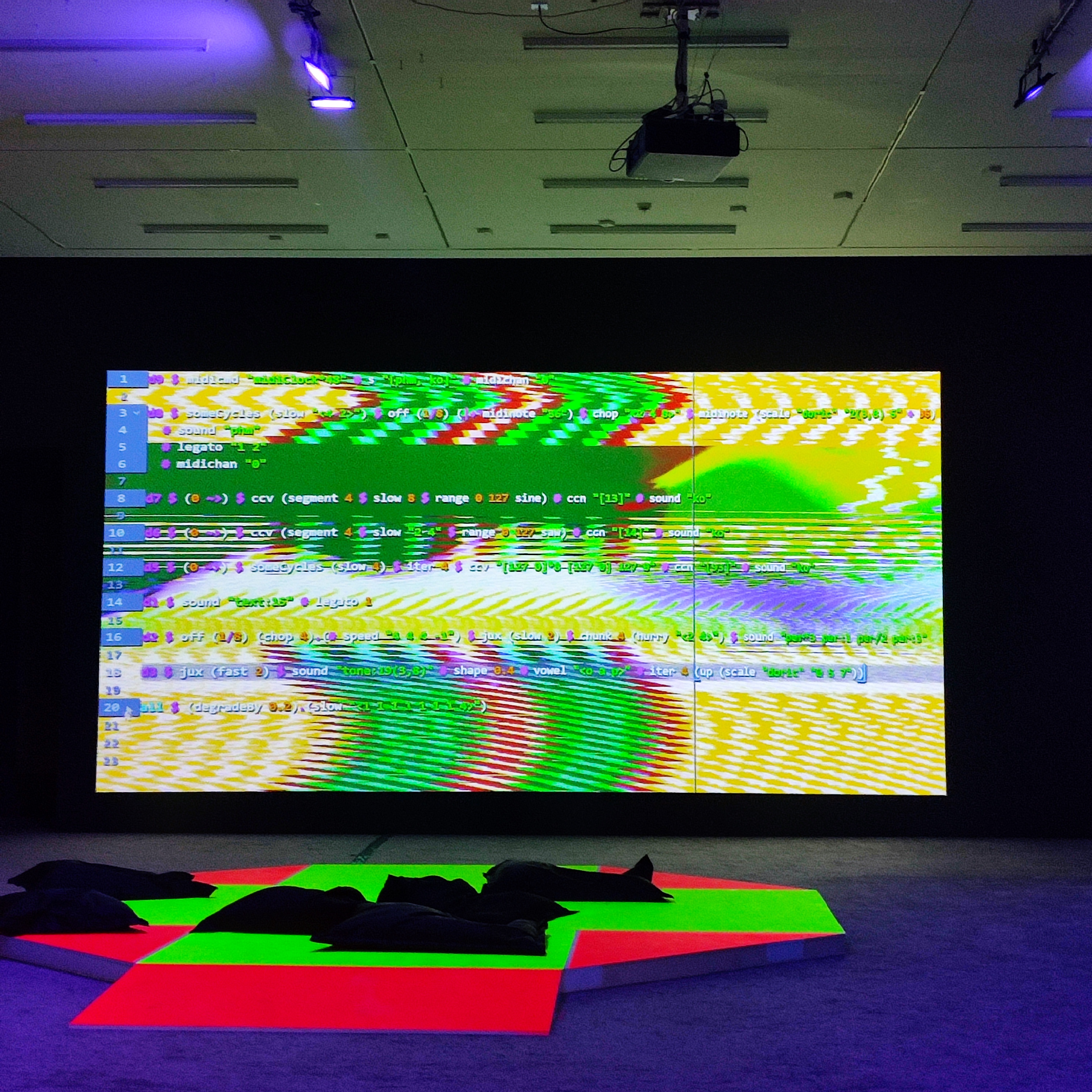 The main piece of the exhibition: a wide projector screen of a video of continuously-typing Strudel code with animation and neon graphics in the background. In front of the screen is a neon red and green raised platform with black pillows for people to sit.